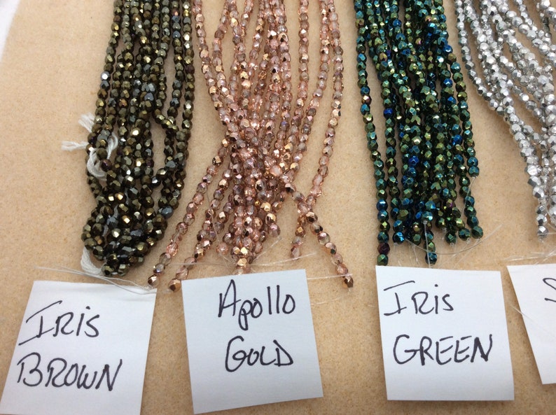 May include: Four strands of glass beads in different colors. The beads are iridescent and have a faceted finish. The strands are labeled "Iris Brown", "Apollo Gold", "Iris Green", and "Silver".