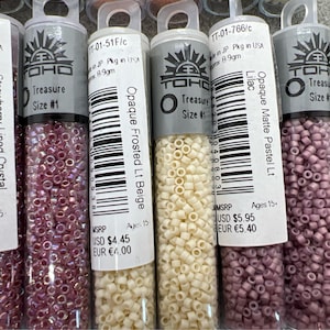 May include: A close-up shot of several small, clear plastic tubes filled with colourful seed beads. The tubes have labels with text including "Strawberry-Lined Crystal Rainbow", "Opaque Frosted Lt Beige", and "Opaque Matte Pastel Lt Lilac". The beads are various shades of pink, beige, and purple.
