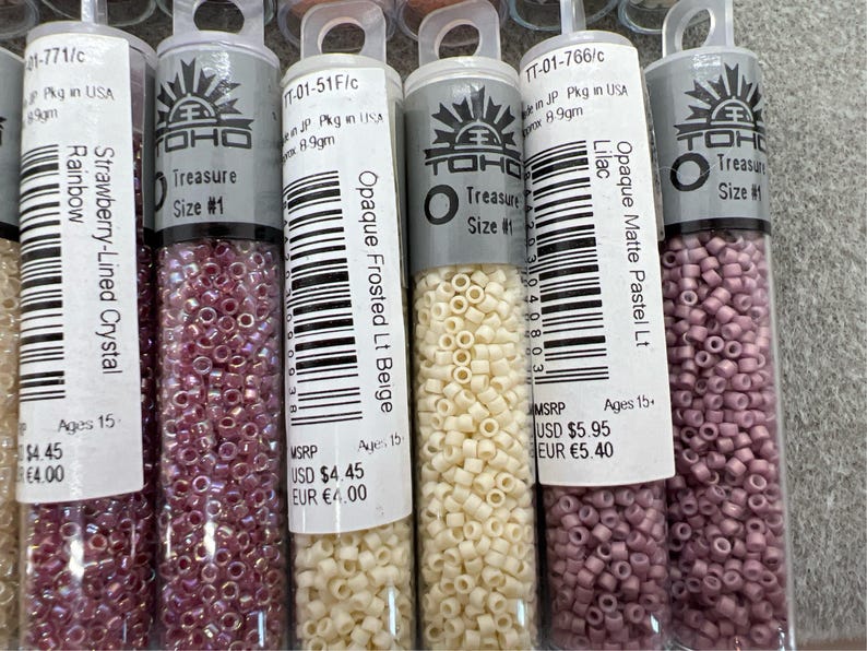 May include: A close-up shot of several small, clear plastic tubes filled with colourful seed beads. The tubes have labels with text including "Strawberry-Lined Crystal Rainbow", "Opaque Frosted Lt Beige", and "Opaque Matte Pastel Lt Lilac". The beads are various shades of pink, beige, and purple.