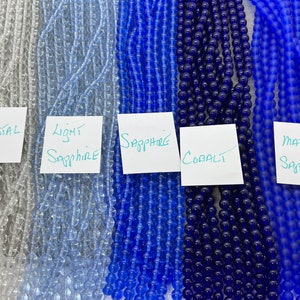 May include: Five strands of glass beads in various shades of blue, from clear to dark blue. The strands are labelled "Crystal", "Light Sapphire", "Sapphire", "Cobalt", and "Matte Sapphire".