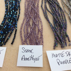 May include: Five strands of glass beads in different colors. From left to right: Jet AB, Stone Amethyst, Matte Iris Purple, Light Grey, and White.