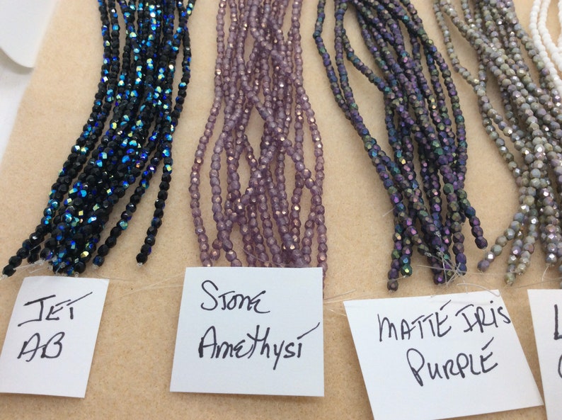 May include: Five strands of glass beads in different colors. From left to right: Jet AB, Stone Amethyst, Matte Iris Purple, Light Grey, and White.