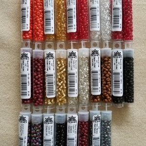 May include: A collection of small, colorful glass beads in clear plastic tubes. The beads come in various colors, including red, gold, silver, and black. Each tube is labeled with product information. These beads are ideal for jewelry making and crafting.
