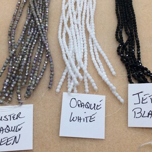 May include: Three strands of glass beads, each with a different color and finish. The left strand is a luster opaque green, the middle strand is opaque white, and the right strand is jet black.