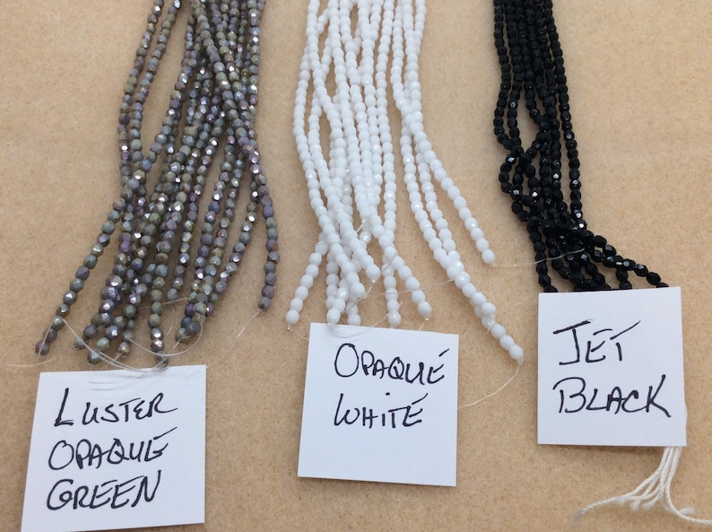 May include: Three strands of glass beads, each with a different color and finish. The left strand is a luster opaque green, the middle strand is opaque white, and the right strand is jet black.