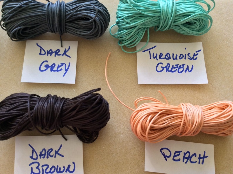 May include: Four spools of waxed cotton cord in different colors: dark grey, dark brown, turquoise green, and peach. The spools are labeled with their respective colors.