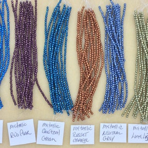 May include: A collection of metallic beads in various colors, including blue, purple, teal, orange, gray, and green. The beads are arranged in rows on a white surface with labels identifying each color.
