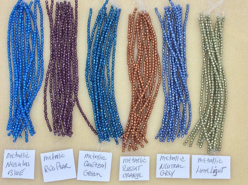 May include: A collection of metallic beads in various colors, including blue, purple, teal, orange, gray, and green. The beads are arranged in rows on a white surface with labels identifying each color.