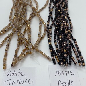May include: Two strands of glass beads. One strand is white with gold flecks and labeled "White Tortoise Celsian". The other strand is black with gold flecks and labeled "Matte Apollo Jet".