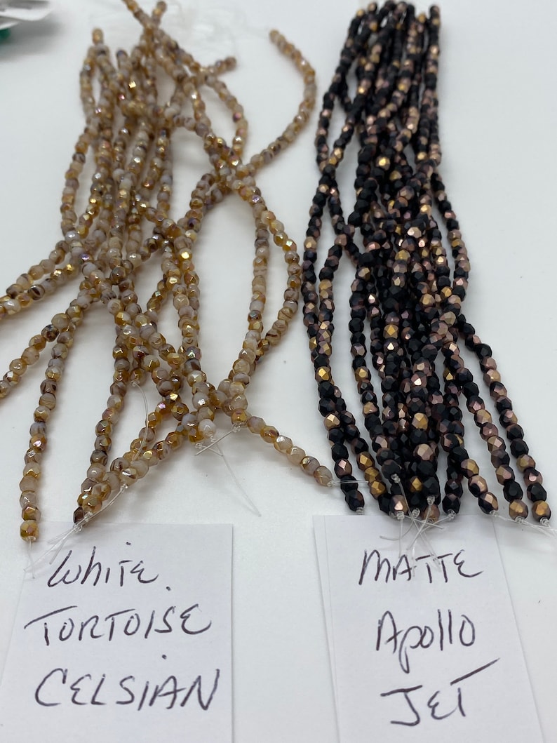May include: Two strands of glass beads. One strand is white with gold flecks and labeled "White Tortoise Celsian". The other strand is black with gold flecks and labeled "Matte Apollo Jet".