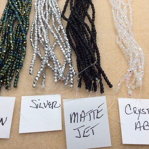 May include: Five strands of beads in different colors and finishes. From left to right: rose gold, iridescent green, silver, matte jet black, and crystal AB. Each strand is labeled with its color and finish.