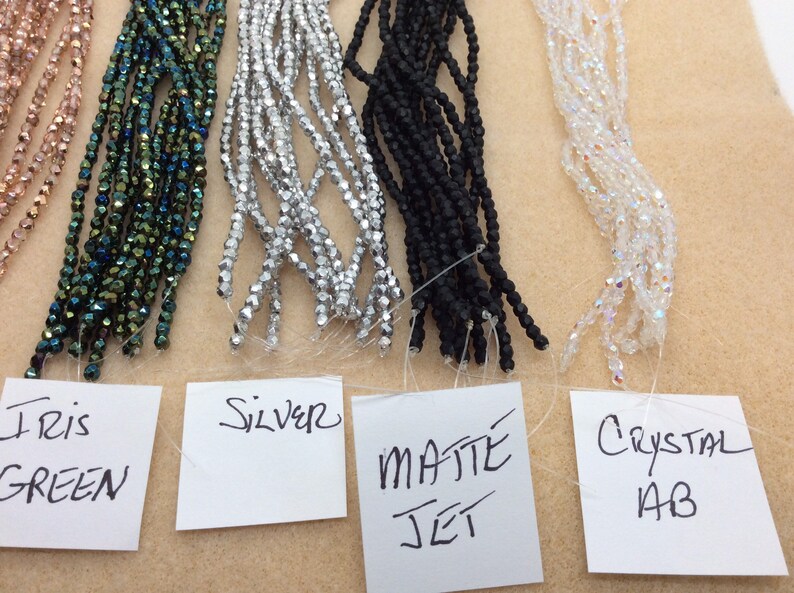 May include: Five strands of beads in different colors and finishes. From left to right: rose gold, iridescent green, silver, matte jet black, and crystal AB. Each strand is labeled with its color and finish.