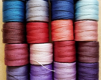 Puede incluir: Una colección de 24 carretes de hilo de bordar en varios colores, incluyendo azul, rojo, morado y blanco. Los carretes están dispuestos en cuatro filas de seis.