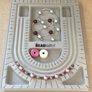 May include: A gray bead board with measurements and the text "The Beadsmith". The board holds several beaded bracelets with pearls and beads in shades of pink, silver, and burgundy. Two spools of thread are also visible.