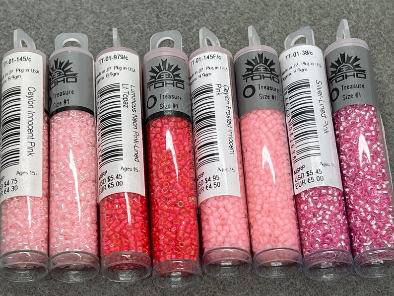May include: Six clear tubes filled with small, colourful beads. The tubes are labelled with the brand "TOHO Treasure Size #1" and various shades of pink, including "Ceylon Innocent Pink" and "Silver-Lined Pink".
