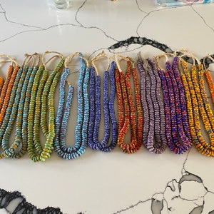 Może przedstawiać: Kolekcja dziesięciu kolorowych naszyjników z koralików, każdy z innym wzorem i połączeniem kolorów. Naszyjniki wykonane są z małych, okrągłych koralików i nawleczone na cienki sznurek. Koraliki są w odcieniach pomarańczowego, zielonego, niebieskiego, fioletowego i żółtego.