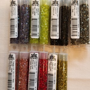 May include: Ten tubes of TOHO seed beads, each containing a different color and finish. The beads are arranged in two rows of five. The tubes are labeled with the bead size, color, and finish. The beads are all 11/0 size and 2.5 inches long.