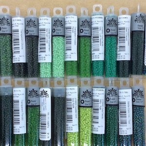 May include: A collection of 11 seed beads in various shades of green, including Milky Greenery, Transparent Olivine, Sea Foam-Lined, Inside Color Crystal/leon, Transparent Frosted, Green Emerald, Milky Lush Meadow, Sour Apple, HYBRID Sueded Gold, Milky-Kale, and Mame-Color ins Teal. The beads are packaged in clear plastic tubes with labels that include the color name, size, and brand.