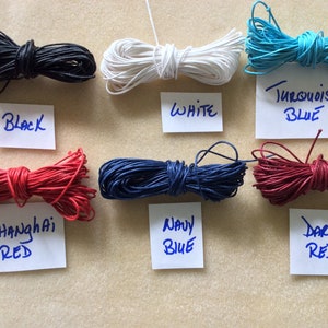 May include: Assortment of waxed cotton cords in various colors, including black, white, navy blue, turquoise blue, red, and dark red. The cords are arranged in rows on a light brown surface. Each cord has a label with the color name written on it.