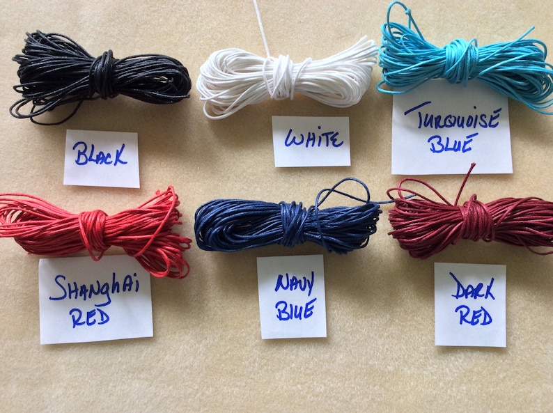 May include: Assortment of waxed cotton cords in various colors, including black, white, navy blue, turquoise blue, red, and dark red. The cords are arranged in rows on a light brown surface. Each cord has a label with the color name written on it.