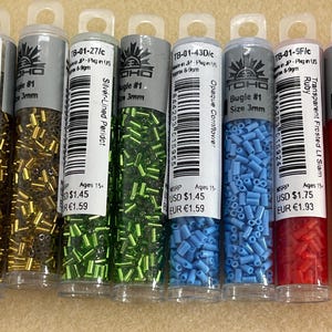 May include: A collection of six clear plastic tubes filled with small, colorful bugle beads. The tubes are labeled with product information, including color names like Copper-Lined Lime Green and Ruby. Each tube contains 3mm beads.
