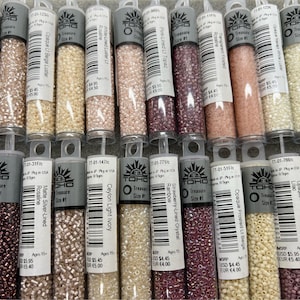 May include: A selection of small glass bead tubes in shades of pink, beige, and purple. Each tube is labelled with product details. The beads are likely for jewellery making or crafts. The tubes are neatly arranged.