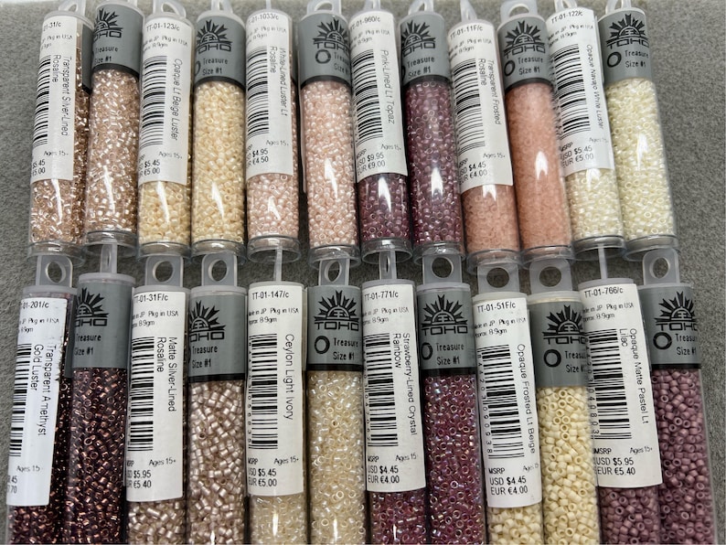 May include: A selection of small glass bead tubes in shades of pink, beige, and purple. Each tube is labelled with product details. The beads are likely for jewellery making or crafts. The tubes are neatly arranged.