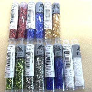 May include: A collection of Toho bugle beads in clear plastic tubes. The beads come in various colors, including red, blue, gold, green, and white. Each tube is labeled with the bead type and size, with some measuring 3mm.
