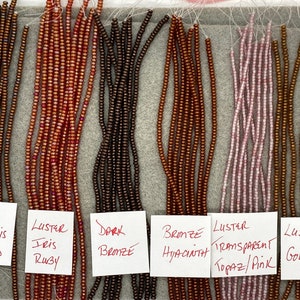 May include: An assortment of seed beads in various colors, including bronze, ruby, and pink, are displayed. Each strand is labeled with its color and finish, such as "Luster Iris" and "Transparent Topaz/Pink". The beads are strung on thin threads.