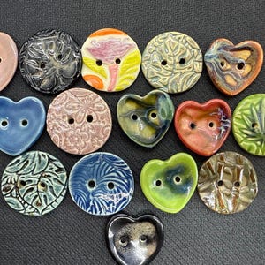 May include: A collection of ceramic buttons in various shapes and colors. The buttons include heart and round shapes, with designs such as floral patterns and leaf motifs. Colors include pink, blue, green, and brown.