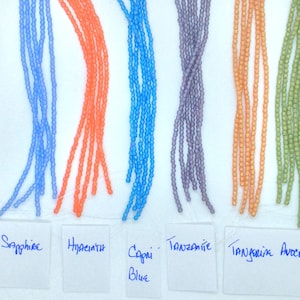 May include: A close-up of ten strands of glass beads in various colors, including red, blue, orange, purple, and green. The beads are arranged in rows on a white surface with labels underneath each row identifying the colors as Siam Ruby, Sapphire, Hyacinth, Capri Blue, Tanzanite, Tangerine, and Avocado.