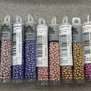 May include: Ten small containers of TOHO brand size 8 round seed beads. The beads are in various colors, including lilac, violet, blush, golden fleece, and sugar plum. Each container has a label with the color name, size, and material.