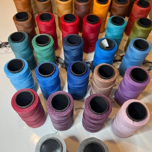May include: A collection of spools of colorful thread in various shades, including red, blue, green, and brown. The spools are cylindrical with black centers, arranged on a light-colored surface. The threads appear to be tightly wound and ready for use.