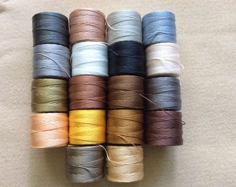 Puede incluir: Una colección de 18 carretes de hilo en varios colores, incluyendo beige, marrón, gris, blanco y amarillo. Los carretes están dispuestos en un patrón rectangular.