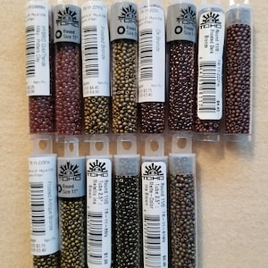 May include: A collection of TOHO brand glass seed beads in clear plastic tubes. The tubes contain various colors, including bronze, brown, and iridescent shades. The labels indicate sizes and finishes like "Frosted" and "Metallic".
