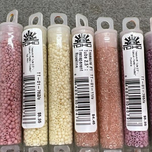 May include: A selection of small, cylindrical beads in clear plastic tubes. The beads are various shades of pink, cream, and peach. Each tube has a label with text including "Treasure #1 Tube 2.5" and a product code. The tubes are arranged in a row.