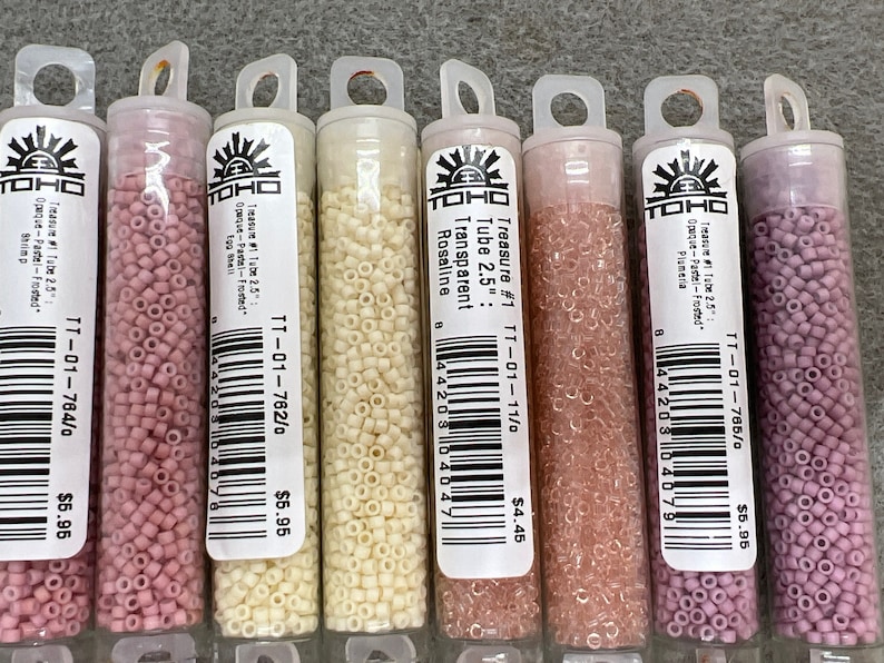 May include: A selection of small, cylindrical beads in clear plastic tubes. The beads are various shades of pink, cream, and peach. Each tube has a label with text including "Treasure #1 Tube 2.5" and a product code. The tubes are arranged in a row.