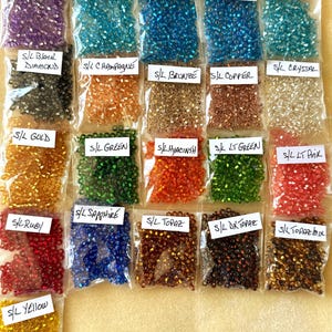 May include: A collection of small, colorful beads in clear plastic bags. Each bag is labeled with a color name, including amethyst, aqua, black diamond, champagne, bronze, copper, crystal, gold, green, hyacinth, lt green, lt pink, ruby, sapphire, topaz, dk topaz, topaz mix, and yellow.