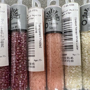 May include: Image of several clear plastic tubes filled with small, colourful beads. The tubes have labels with text including "Pink-Lined Lt Topaz", "Transparent Frosted Rosaline", and "Opaque Navajo White Luster". The beads are various colours.
