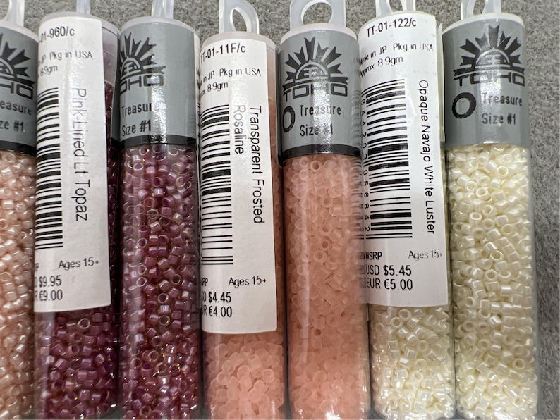 May include: Image of several clear plastic tubes filled with small, colourful beads. The tubes have labels with text including "Pink-Lined Lt Topaz", "Transparent Frosted Rosaline", and "Opaque Navajo White Luster". The beads are various colours.