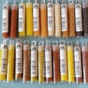 May include: A collection of small, colorful glass beads in clear tubes. The beads are in various shades of yellow, orange, brown, and white. Each tube has a label with text and a price tag.