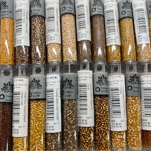 May include: A close-up view of 12 small tubes of Toho Treasure Size #1 beads. The tubes are labeled with different colors and descriptions, including Transparent Marmalade, Yellow-Lined, Frosted Gold-Lined Crystal, Silver-Lined Frosted Onyx, Topaz, and Transparent Topaz.
