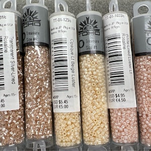 May include: A selection of small glass bead tubes in shades like silver-lined, beige, and pink. Each tube is labelled with product details and the brand logo. The beads are suitable for crafting and jewellery making.