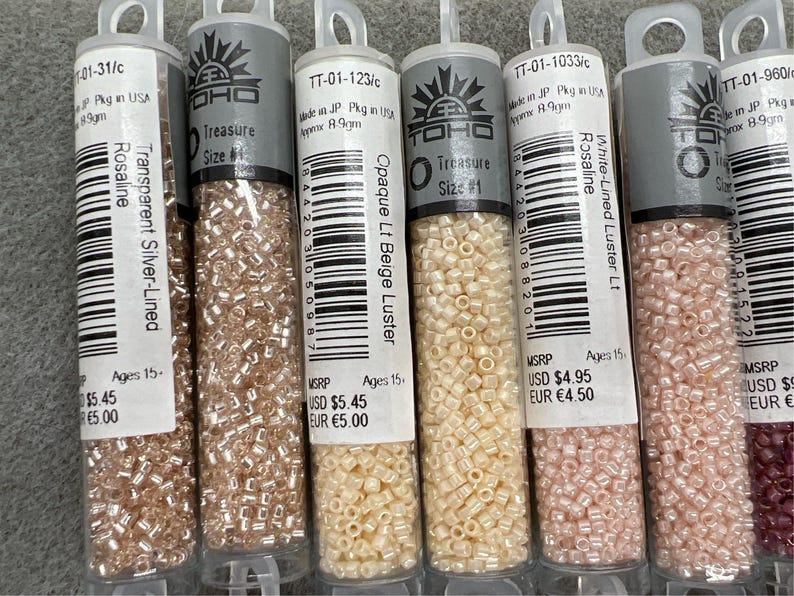May include: A selection of small glass bead tubes in shades like silver-lined, beige, and pink. Each tube is labelled with product details and the brand logo. The beads are suitable for crafting and jewellery making.