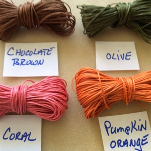 May include: Close-up of five spools of waxed cotton cord in different colors: chocolate brown, olive green, coral pink, pumpkin orange, and teal blue. Each spool is labeled with its color name.