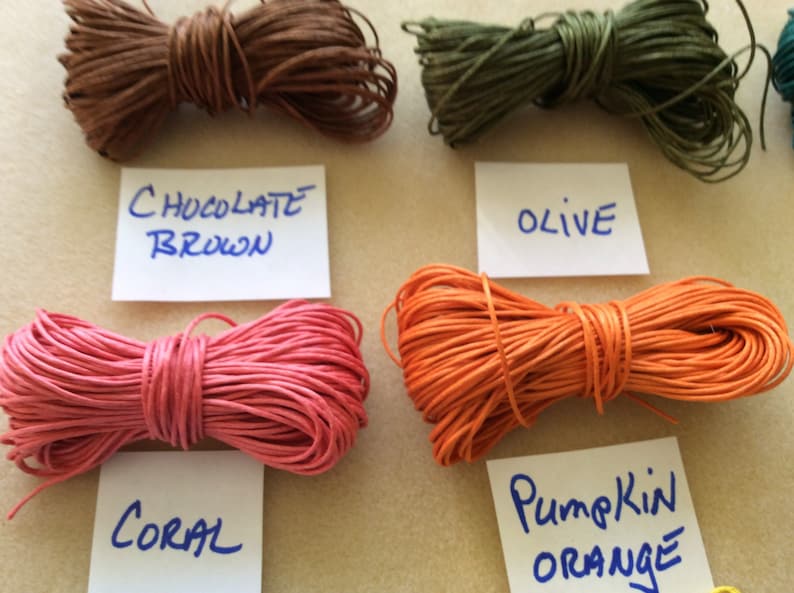 May include: Close-up of five spools of waxed cotton cord in different colors: chocolate brown, olive green, coral pink, pumpkin orange, and teal blue. Each spool is labeled with its color name.