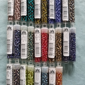 May include: A collection of 20 small tubes of TOHO seed beads. The tubes are arranged in three rows. Each tube contains a different color and type of bead. The beads are round and have a variety of finishes, including metallic, iridescent, and opaque.