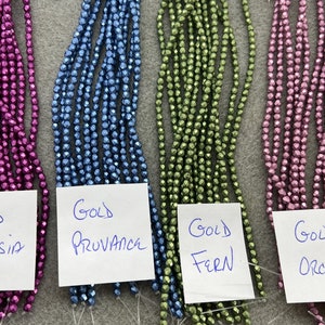 May include: Four strands of faceted metallic beads in fuchsia, blue, green, and pink. Each strand has a white paper label with handwritten text: Gold Fuchsia Red, Gold Provance, Gold Fern, and Gold Orchid, identifying the specific color of each bead.
