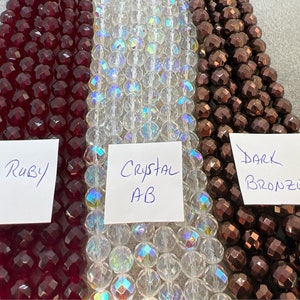 May include: Close-up of three strands of faceted beads in ruby red, crystal AB, and dark bronze colors. The beads are labeled "Ruby", "Crystal AB", and "Dark Bronze" on white paper.