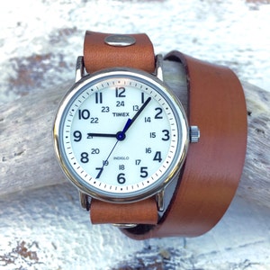 May include: A white analog wristwatch with a brown leather band. The watch face has black hands and black numbers. The watch face also has a blue second hand and the text "TIMEX" and "INDIGLO" printed on it.